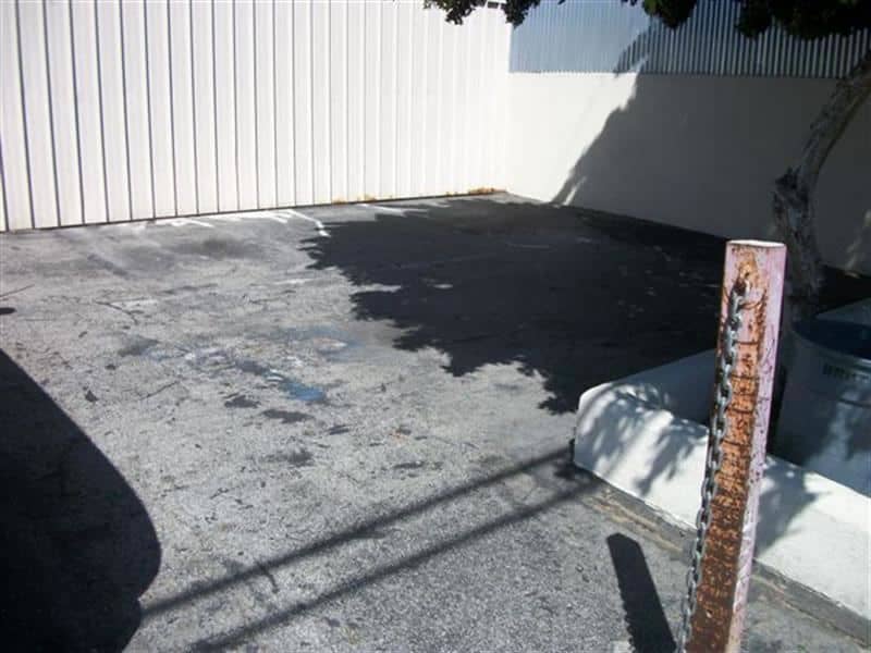 Badly cracked asphalt corner beside a building, with patched areas and broken edges