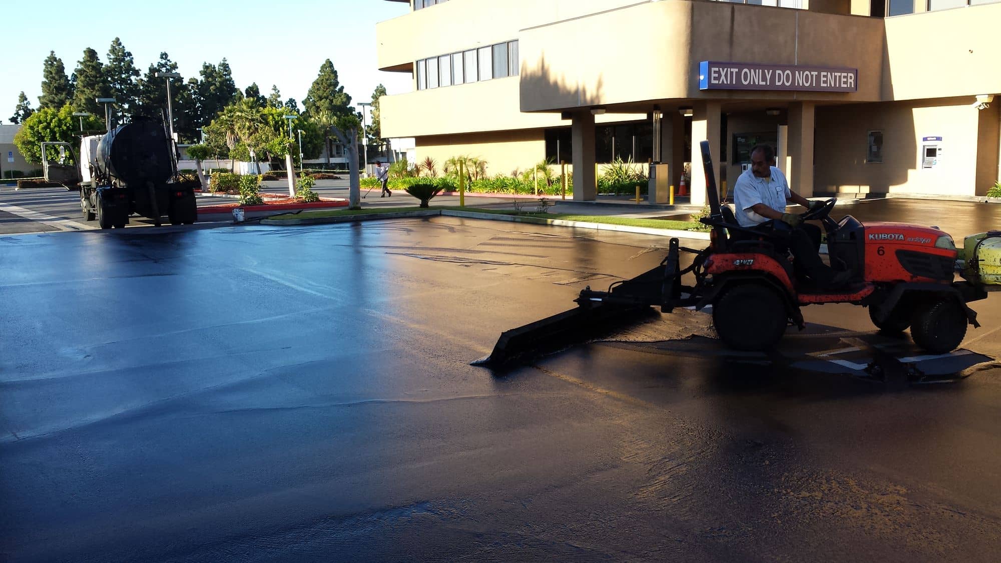 Sealcoat tractor spreading sealer in front of an office building as the morning light hits the wet surface