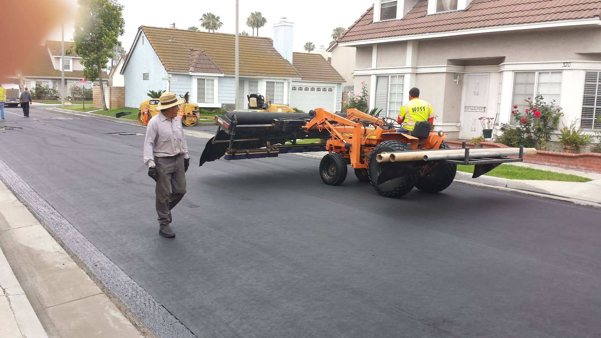 Tractor-drawn rig smoothing paving fabric on a neighborhood street lined with two-story homes