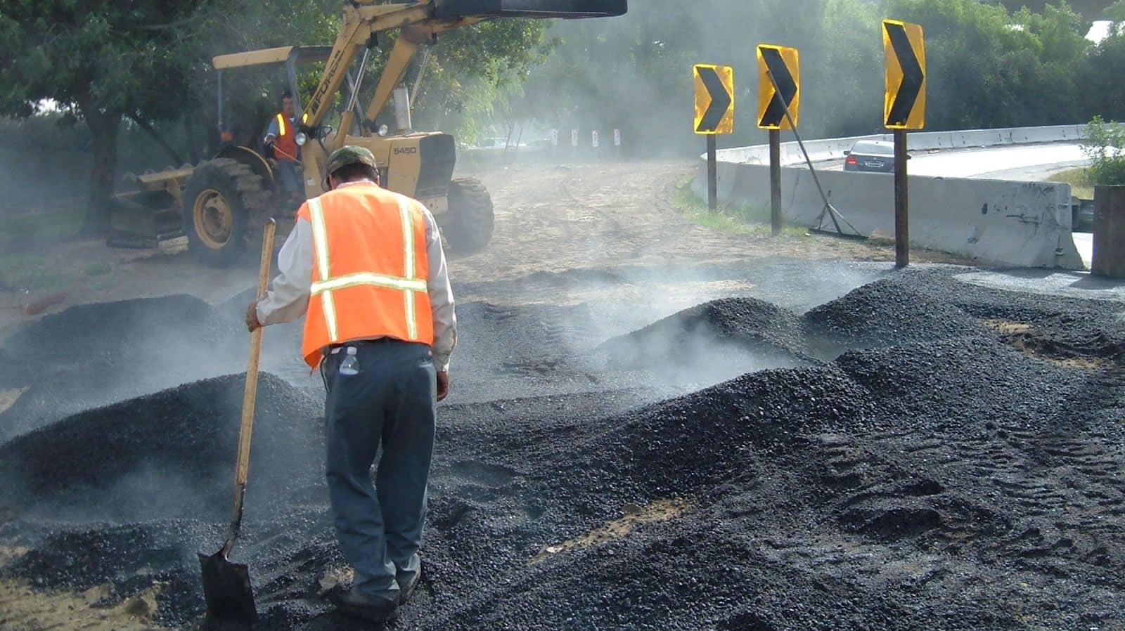 Crew member in a safety vest shoveling steaming asphalt material beside a roadway as a loader works behind