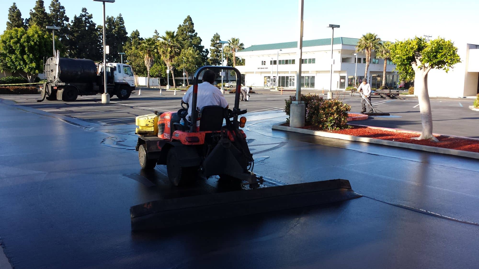 Tractor dragging a wide squeegee across a parking lot, leaving a glossy coat of fresh sealer