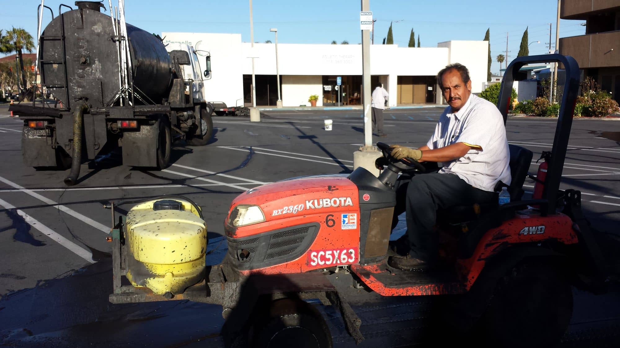 Operator on a compact tractor fitted with a sealcoat spreader, tank truck in the background