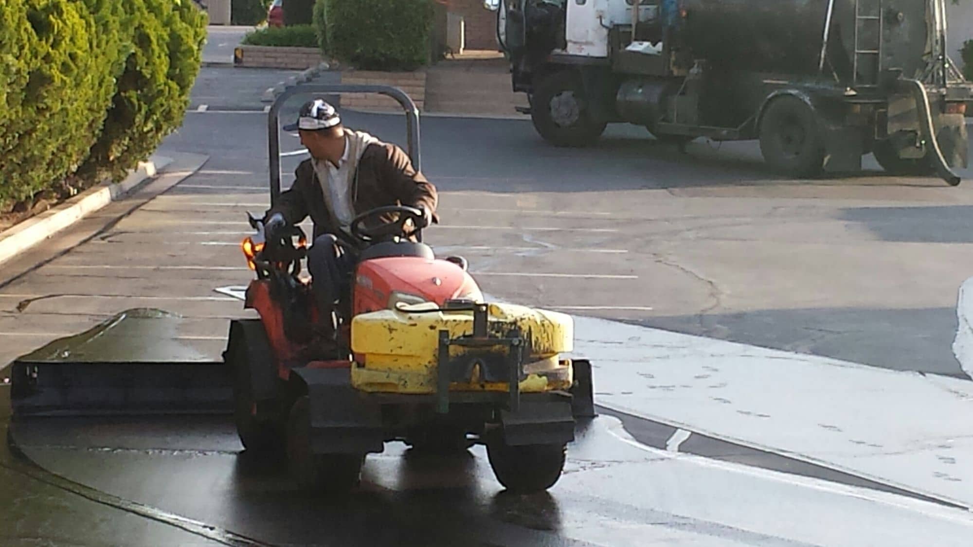 Operator driving a sealcoat tractor across a lot with the tank truck parked behind