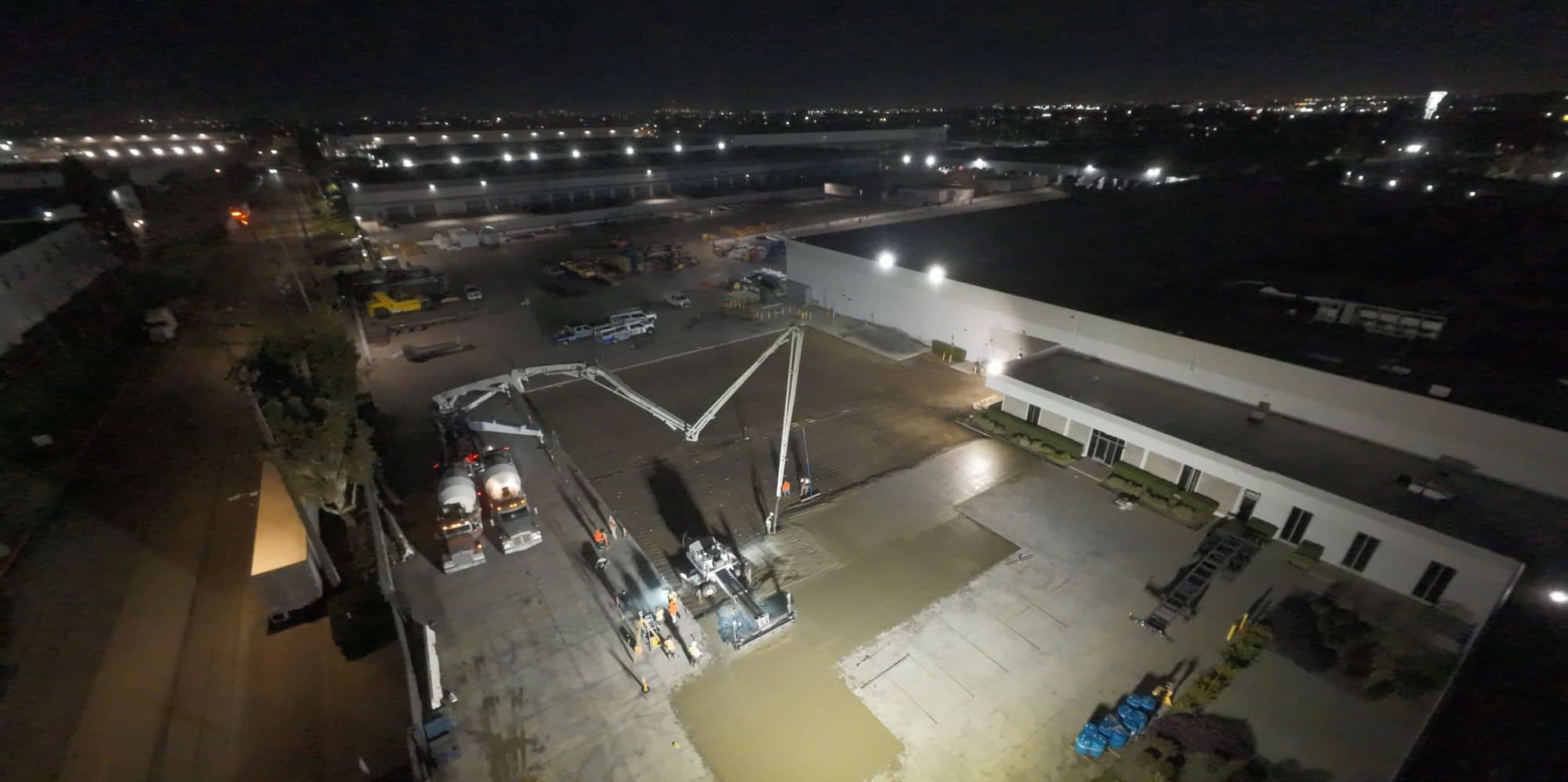 Aerial night view of a large concrete pour beside a warehouse, with pump booms and trucks working under floodlights