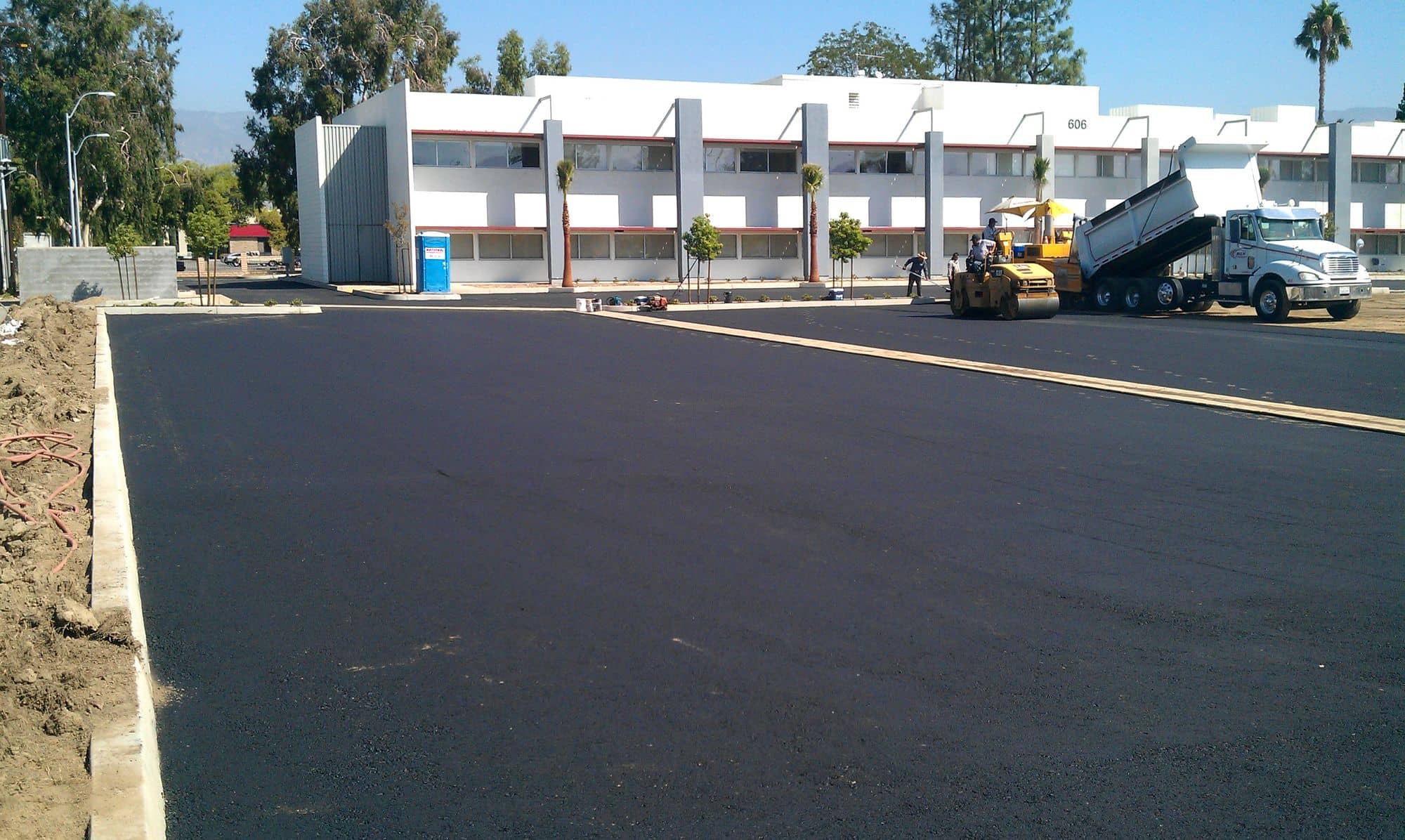 Freshly paved black asphalt lot beside a white commercial building, with a dump truck and loader at the far edge