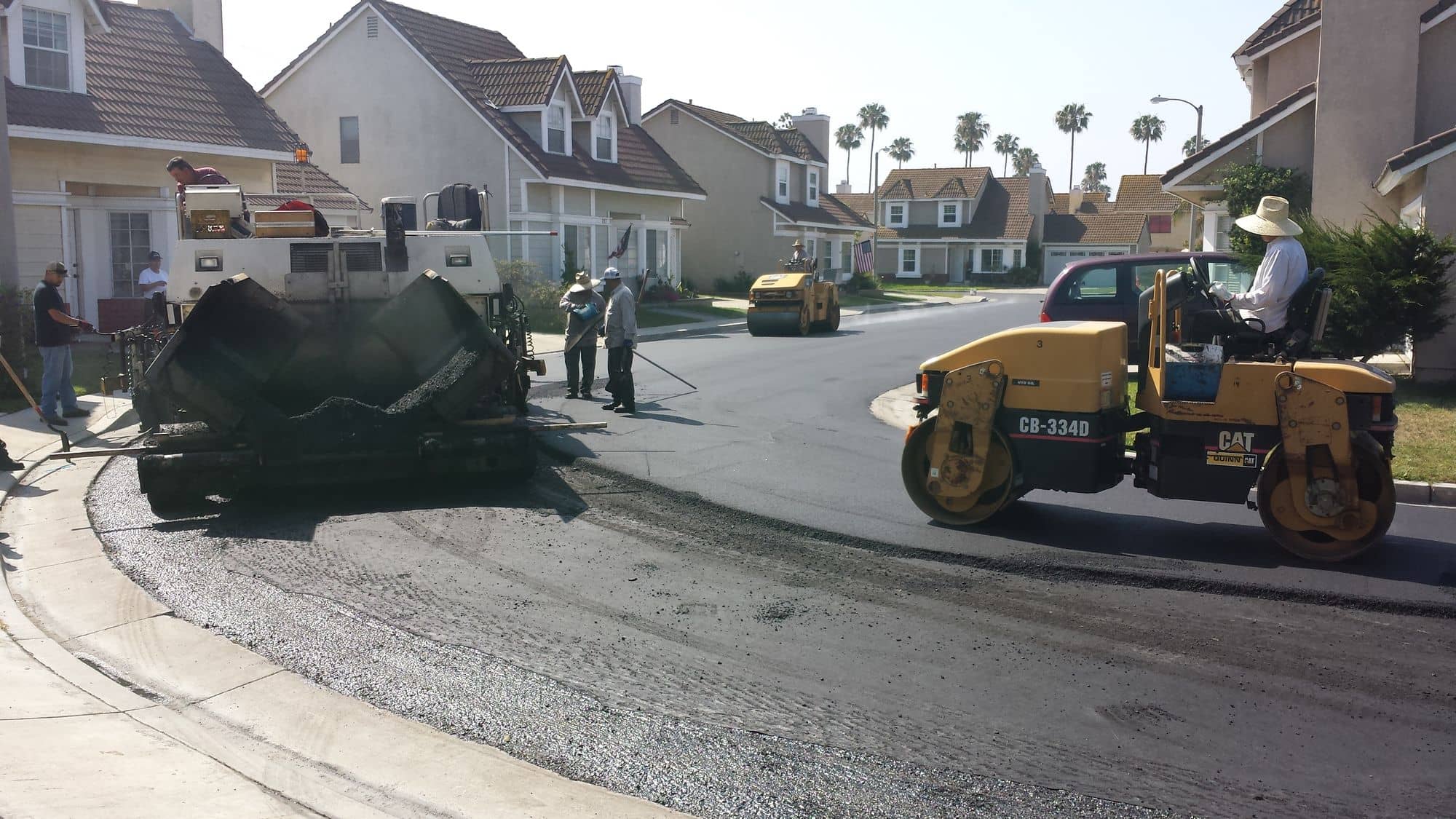 Asphalt paver laying a new mat around a residential corner while a double-drum roller compacts behind it