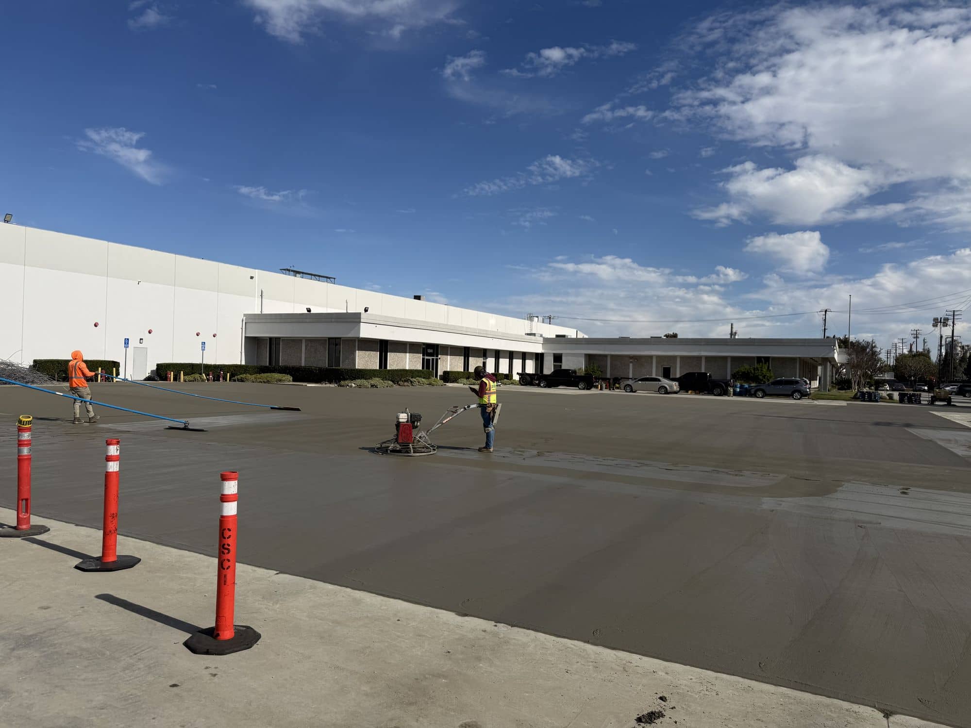 Finishers working across a freshly placed concrete slab outside a warehouse, with delineators along the edge
